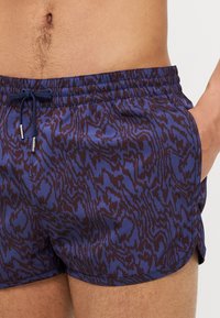 Shorts de bain en violet foncé avec un motif abstrait marron. Dotés d'une taille élastique, d'un cordon de serrage et de poches latérales. Texture lisse.