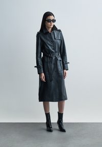 Črni usnjeni dolg plašč z pasom v pasu, zapenjanjem na gumbe in velikimi žepi; kombiniran z elegantnimi črnimi gležnarji in velikimi sončnimi očali.