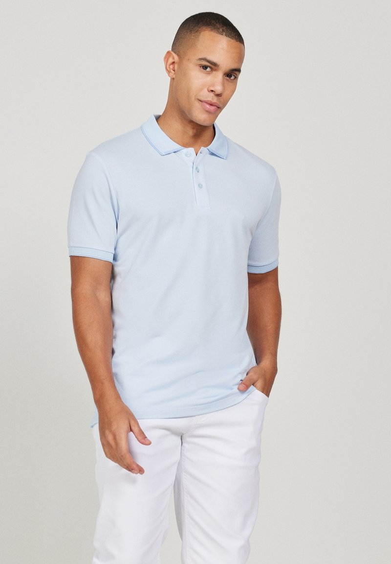 Jeune homme portant un polo bleu clair à manches courtes et un pantalon blanc, debout avec une main dans la poche devant un fond uni.