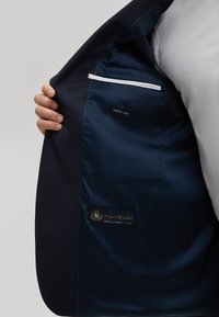 Blazer in lana navy con fodera blu in raso. L'interno presenta un'etichetta che indica "SUPER 130's WOOL" e dettagli tessuti dall'Italia.