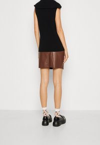 Schwarzes ärmelloses Strickoberteil mit umgeschlagenem Kragen, kombiniert mit einem braunen Leder-Minirock. Getragen mit gemusterten Socken und klobigen schwarzen Schuhen.