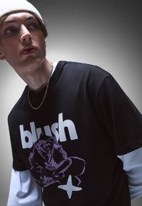 Black t-shirt featuring bold white "blush" text and a purple rose graphic. Layered over a light blue long-sleeve shirt. Silver chain necklace.