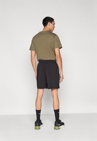 Κοντομάνικο μπλουζάκι ελιάς και μαύρο αθλητικό σορτς με ελαστική μέση και πλαϊνές τσέπες, φορεμένα με μαύρα αθλητικά παπούτσια και κάλτσες.