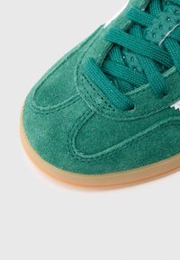 Zöld velúr tornacipő, texturált felsőrésszel, lapos fűzőkkel és gumiból készült külső talppal. A cipő oldalán fehér részletek találhatók.