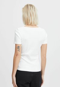 White short-sleeve fitted t-shirt with a round neck, ribbed texture, and a visible tattoo on the left arm. Black jeans are worn below.