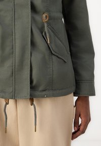 Veste en coton vert olive avec fermeture éclair et bouton pression, une poche à rabat, et des cordons de serrage à la taille et à l'ourlet.