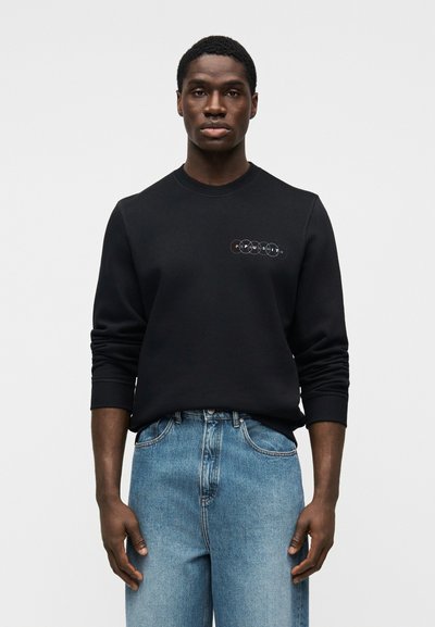 Sweat-shirt noir à col rond, avec une broderie de logo circulaire multicolore sur la poitrine. Associé à un jean en denim bleu clair.