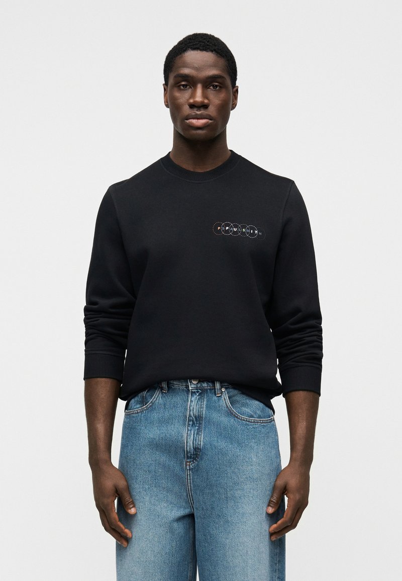 Μαύρο φούτερ με στρογγυλή λαιμόκοψη, με πολύχρωμ ο κεντημένο κυκλικό λογότυπο στο στήθος. Συνδυασμένο με ανοιχτό μπλε τζιν παντελόνι.