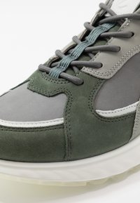 Sneaker in suede verde e grigio con accenti texturizzati, dotata di suola spessa, lacci piatti e design a strati sulla parte superiore.