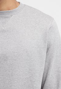 Šedá mikina s žebrovaným kulatým výstřihem a texturovanou tkaninou. Postrádá výrazné vzory nebo zdobení, zaměřuje se na jednoduchost a pohodlí.