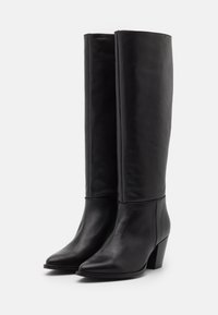 Paire de bottes hautes en cuir noir avec des bouts pointus et des talons carrés, placées côte à côte sur un fond blanc uni.