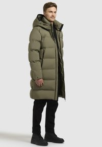 Olivengrøn pufferjakke med lang design, polstret tekstur, stor hætte, fuld frontlynlås og sorte detaljer; båret over sorte bukser og støvler.