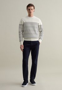 White sweater with navy stripes, round neck, ribbed cuffs; paired with navy trousers and dark sneakers, standing against a neutral background.