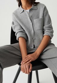 Cardigan gris côtelé avec un col à revers, une poche poitrine, et une fermeture à boutons, porté avec un pantalon gris, assis sur une chaise noire.