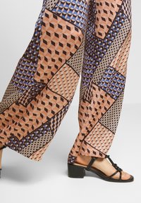 Kalhoty s širokými nohavicemi s geometrickým vzorem v broskvové, modré a hnědé barvě, doplněné černými sandály na řemínky s masivním podpatkem.