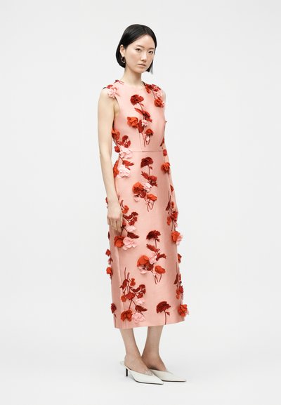 Naine, kes kannab varrukateta roosat midi-pikade punaste ja roosade 3D-lilleaplikatsioonidega kleiti, kombineeritud valgete teravate nina ja madala kontsaga kingadega, seisab külgvaates.