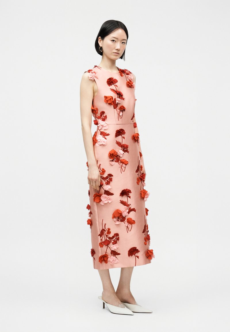 Donna che indossa un vestito midi rosa senza maniche con applicazioni floreali 3D rosse e rosa, abbinato a décolleté bianchi con tacco kitten a punta, in piedi di profilo.