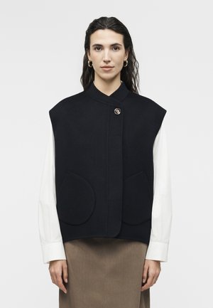 Sötét hajú nő fekete ujjatlan mellényt visel fehér hosszú ujjú ing fölött, bézs szoknyában, egyszínű fehér háttér előtt áll.