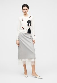 Καρντιγκάν σε λευκό πλεκτό με γραφικό μοτίβο σε μαύρο και κόκκινο, συνδυασμένο με γκρι φούστα με δαντελένιο τελείωμα. Τα παπούτσια είναι μυτερά, εκρού.