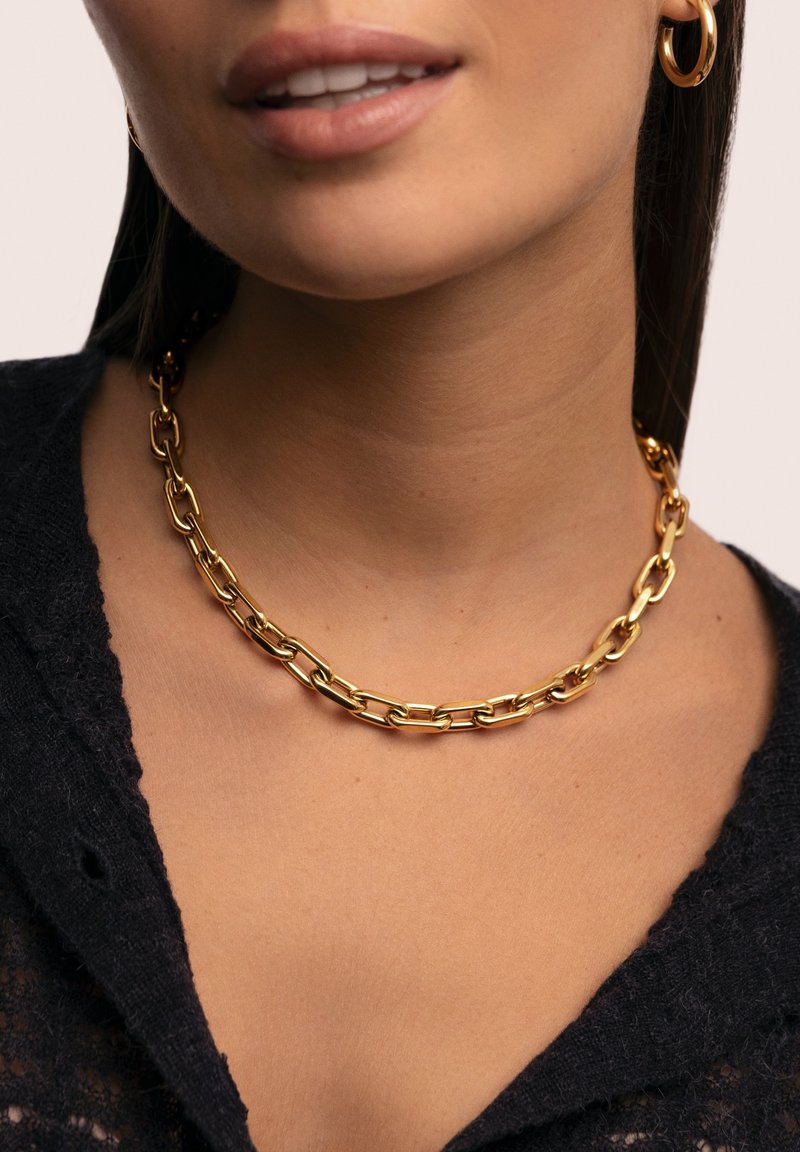 Gros plan d'une femme portant un collier chaîne épais en or et une boucle d'oreille créole assortie, avec un haut texturé noir.