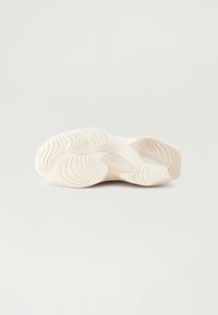 Semelle de basket blanche avec des rainures texturées et ondulées et des accents beiges lisses, vue par le dessous sur un fond uni.