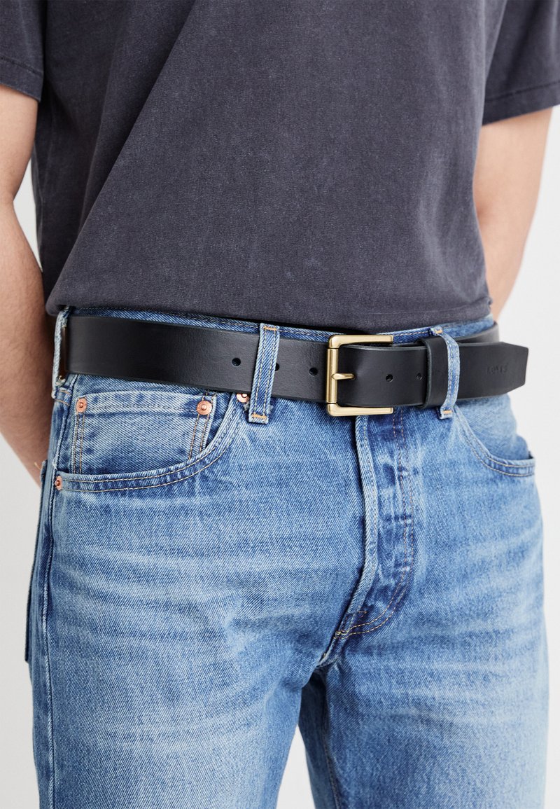 Sort læderbælte med en rektangulær guldmetals spænde, båret med blå denim jeans. Remmen viser flere huller til størrelsesjustering.