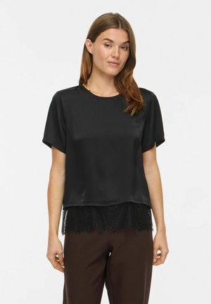 Femme aux cheveux châtain clair portant un haut en satin noir à manches courtes avec bordure en dentelle et pantalon marron foncé sur un fond uni.