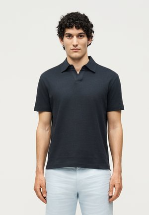 Tânăr cu păr creț, purtând un tricou polo cu mâneci scurte bleumarin închis și pantaloni bleu deschis, stând în fața unui fundal simplu.