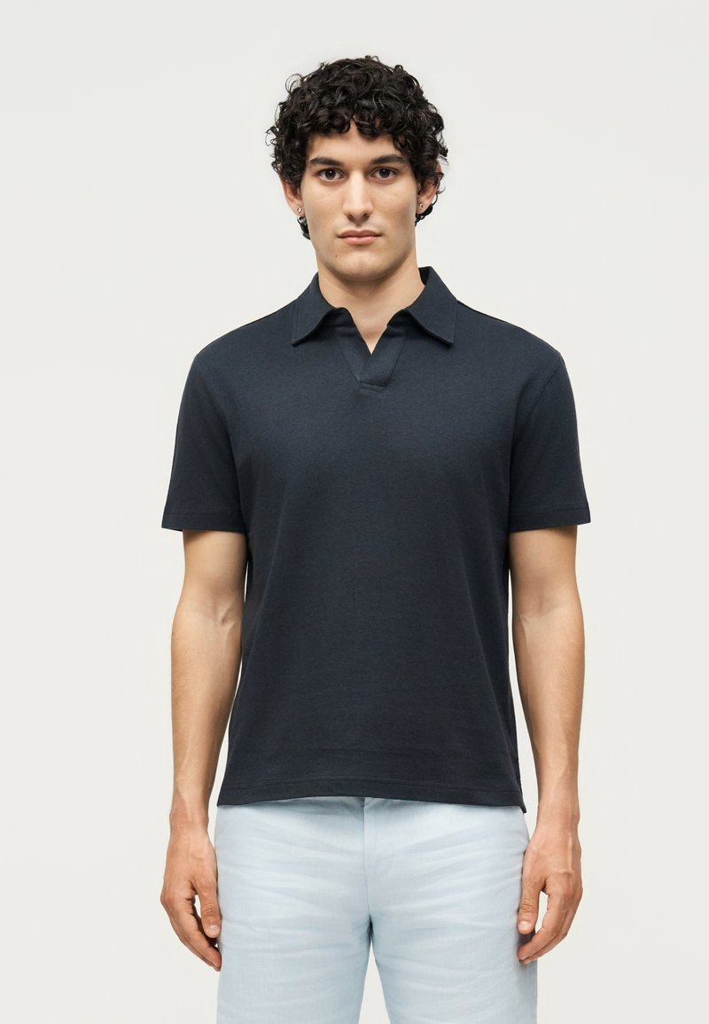 Νεαρός άνδρας με κατσαρά μαλλιά, φοράει σκουρόχρωμο μπλε κοντομάνικο πόλο και ανοιχτό μπλε παντελόνι, στέκεται μπροστά σε απλό φόντο.