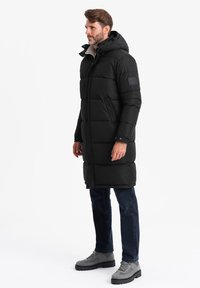 Sort puffertjakke med hætte, med kviltet mønster, frontlynlås og justerbare manchetter. Bæres over mørke jeans og grå støvler.