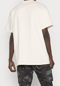Cream-colored cotton t-shirt with a relaxed fit. Short sleeves and a round neckline. Worn with dark camo-patterned cargo pants.