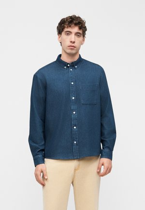 Jeune homme portant une chemise en jean bleu foncé boutonnée avec poche poitrine et un pantalon beige, debout devant un fond blanc.
