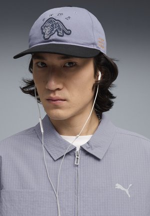 Jongere draagt een paarse Puma-pet met een zwarte klep, een bijpassend ritsjack en witte bedrade oordopjes tegen een grijze achtergrond.