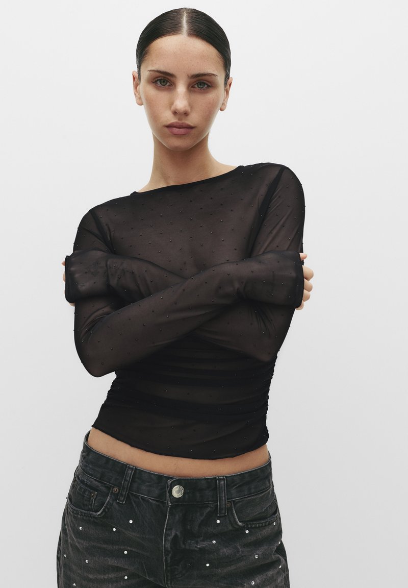 Bluză neagră transparentă cu mâneci lungi, cu un model punctat, textură moale și un design cu pliu, asociată cu blugi închisi la culoare cu broderii.