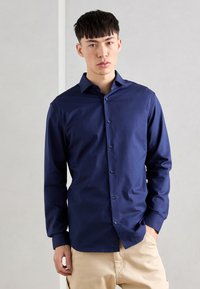 Cămașă navy blue cu mânecă lungă și butoane, cu guler, având butoane pe față și o textură netedă. Poartă cu pantaloni bej.