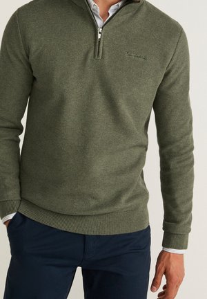 Homme portant un pull texturé olive à fermeture éclair au quart, par-dessus un pantalon bleu et une chemise à col rayée claire.