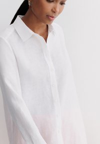 Camicia di lino bianca con un sottile gradiente rosa all'orlo. Presenta un colletto classico e una chiusura con bottoni, con maniche lunghe.
