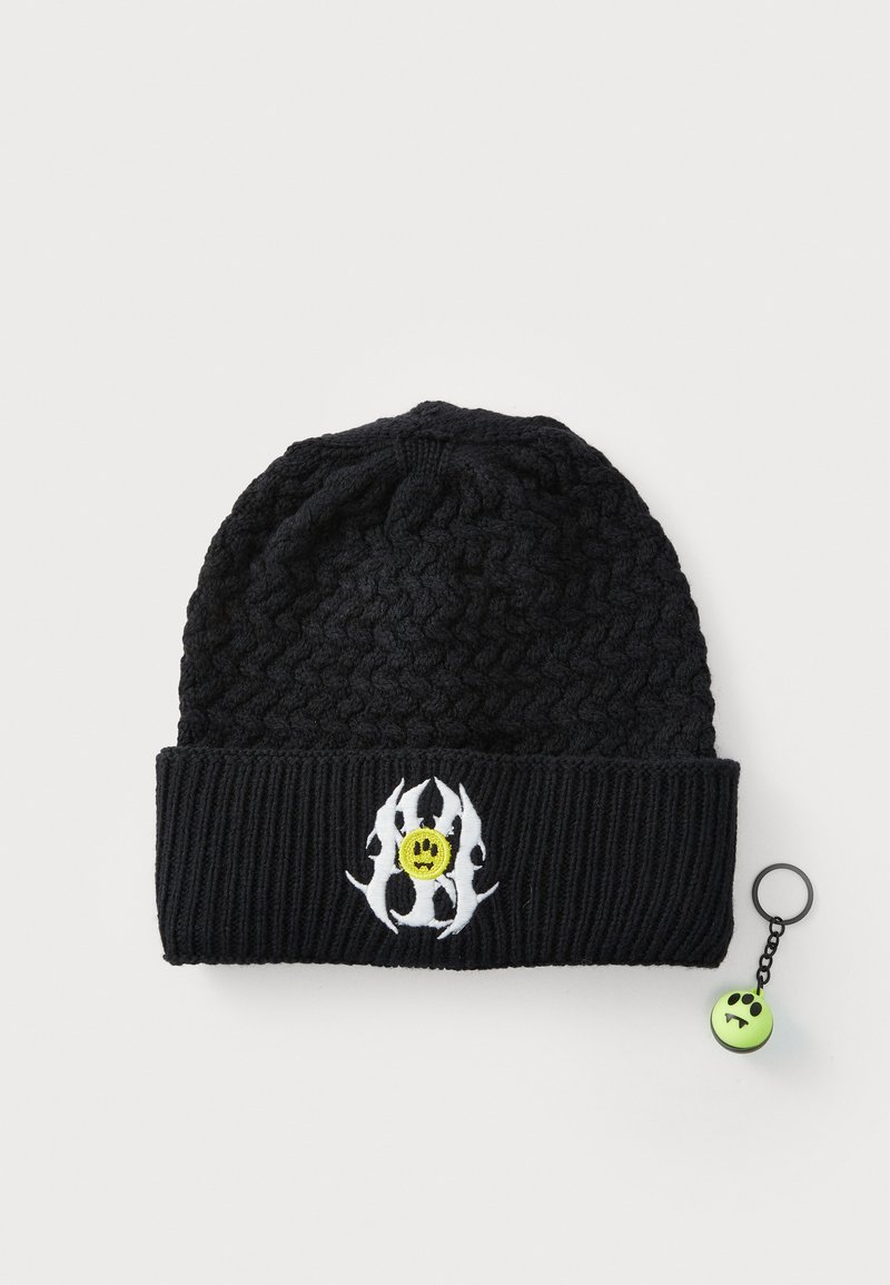 Caciulă tricotată neagră cu manșetă ribbată, având un design cu flăcări albe și o față zâmbitoare galbenă. Include un accesoriu cheie sub formă de față zâmbitoare verde.