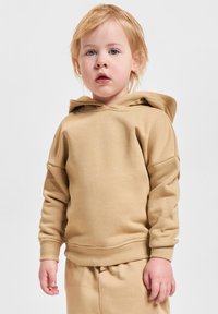 Niño pequeño con cabello claro, vistiendo una sudadera beige y pantalones a juego, de pie contra un fondo claro y liso.
