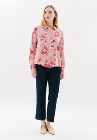Donna che indossa una blusa a righe floreali rossa e bianca, jeans cropped blu scuro e ballerine beige a punta, in piedi davanti a uno sfondo bianco semplice.