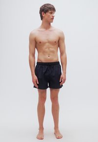 Μαύρα μαγιό σορτς με ελαστική μέση με κορδόνι, λεία υφή και κλασικό κόψιμο, που φοριούνται από έναν μοντέλο χωρίς πουκάμισο που στέκεται ξυπόλητος.