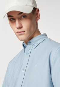 Chemise bleu clair à manches longues avec un col structuré et un logo brodé. Casquette blanche portée légèrement de côté, mettant en valeur la texture de la chemise.