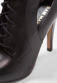 Musta nahkainen korkokengän, jossa on nyöritys, sileä pinta, kärkiosa ja hoikka piikkikorko. Kultainen logo kengän sisäpuolella.