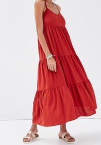 Robe longue rouge sans manches avec un design à étages, présentant un décolleté en V et un tissu fluide. Portée avec des sandales plates de couleur claire.