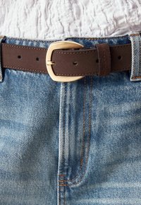 Ceinture en cuir marron avec une boucle en métal doré, portée sur un jean en denim bleu avec des coutures orange et un haut blanc texturé.
