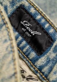 Schwarzes Stofflabel mit weißer "Sheedl est. 1997" Stickerei auf blauem Denim, Nahaufnahme von gelben Nähnähten und Stoffstruktur.