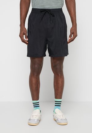 Modelo masculino con pantalones cortos negros con cordón, calcetines a rayas en azul claro y verde, y zapatillas blancas, posando frente a un fondo blanco liso.