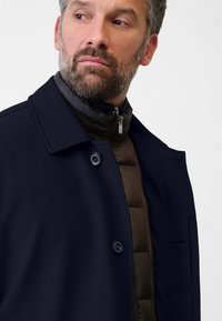 Mörkblå kappa med krage, knäppt fram, lager på en brun puffjacka. Har slät tyg och välstrukturerad design.