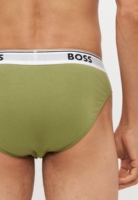 Zöld pamutszabadidő nadrág, fehér elasztikus derékpánttal, rajta a "BOSS" márkanevével. A dizájn szoros illeszkedésű, sima szélekkel és minimális varratokkal rendelkezik.