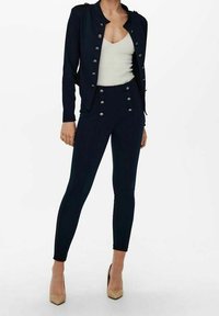 Veste sur mesure bleu marine avec boutons argentés, associée à un haut crème et un pantalon bleu marine ajusté, complétée par des chaussures à talons hauts couleur nude.